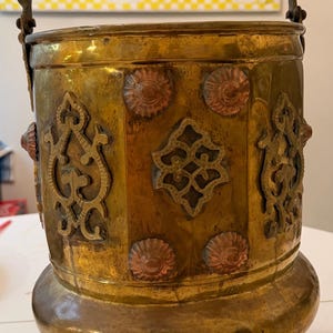 May include: An antique brass bucket with a decorative handle and ornate detailing. The bucket features copper accents and intricate patterns, standing on three small feet. The overall color is a warm, aged brass.