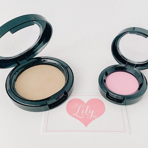 Könnte beinhalten: Zwei runde Make-up-Kompaktverpackungen mit Spiegeln. Die größere Verpackung enthält ein hellbeiges Puder und die kleinere ein hellrosa Puder. Beide Verpackungen sind schwarz mit silbernem Rand.