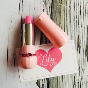 May include: Pink lipstick in a pink and gold tube with a bow. The tube is open and the lipstick is visible. The lipstick is on a white card with the text "Little Lily Shop" in pink.