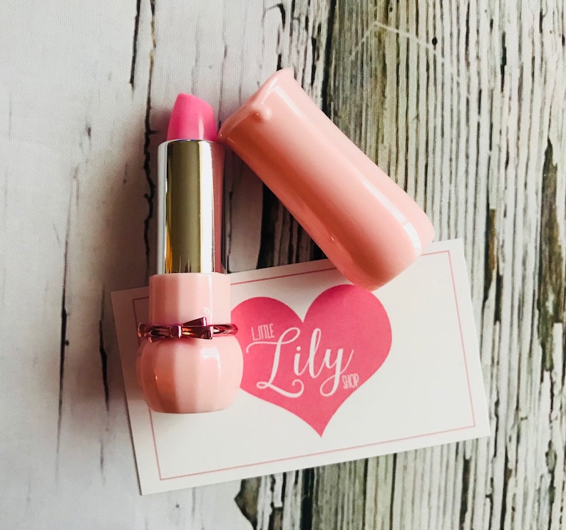 May include: Pink lipstick in a pink and gold tube with a bow. The tube is open and the lipstick is visible. The lipstick is on a white card with the text "Little Lily Shop" in pink.