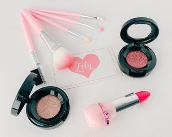 Op de afbeelding: Een set roze make-upkwasten, een roze hartvormige kaart met de tekst "Little Lily", twee zwarte ronde oogschaduwpaletten met roze glinsterende oogschaduw en een roze lipstick met een rode punt.