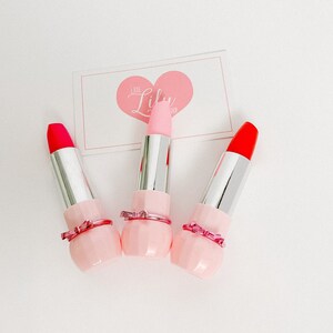 May include: Three pink lipstick tubes with a pink bow detail. The lipsticks are red, pink, and orange.