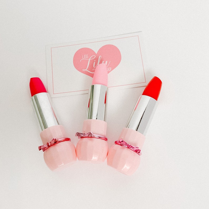May include: Three pink lipstick tubes with a pink bow detail. The lipsticks are red, pink, and orange.