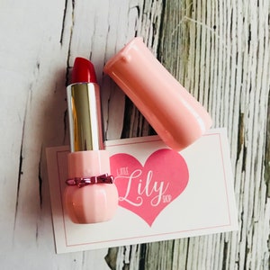 May include: A pink lipstick tube with a red lipstick bullet and a pink bow. The tube has a heart-shaped design with the text "Little Lily Shop" on it.