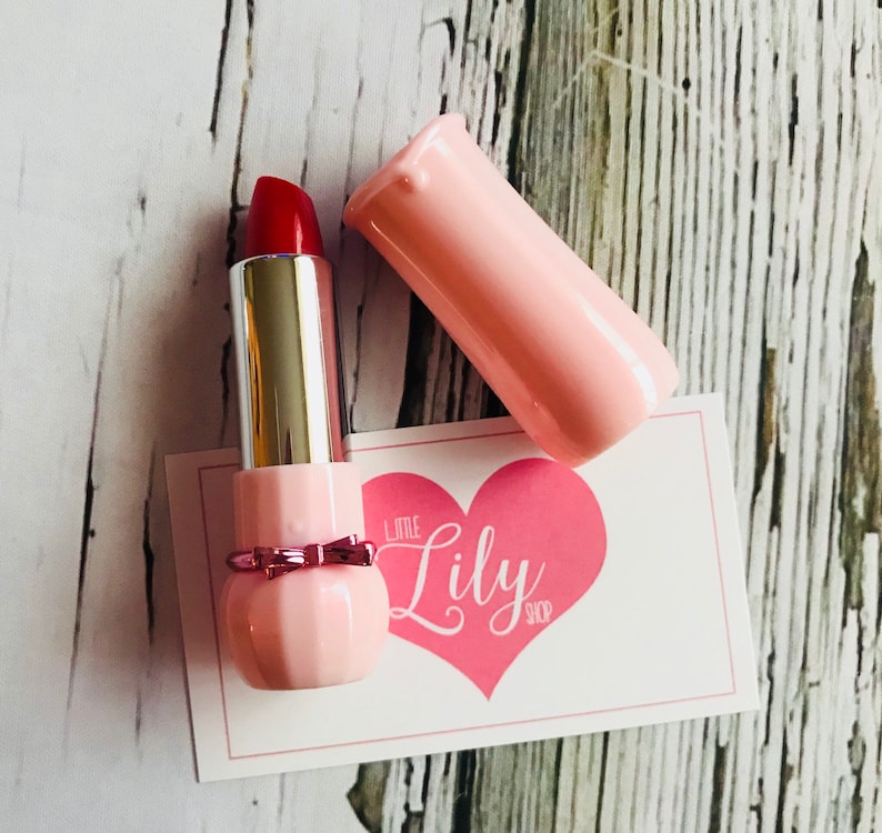 May include: A pink lipstick tube with a red lipstick bullet and a pink bow. The tube has a heart-shaped design with the text "Little Lily Shop" on it.