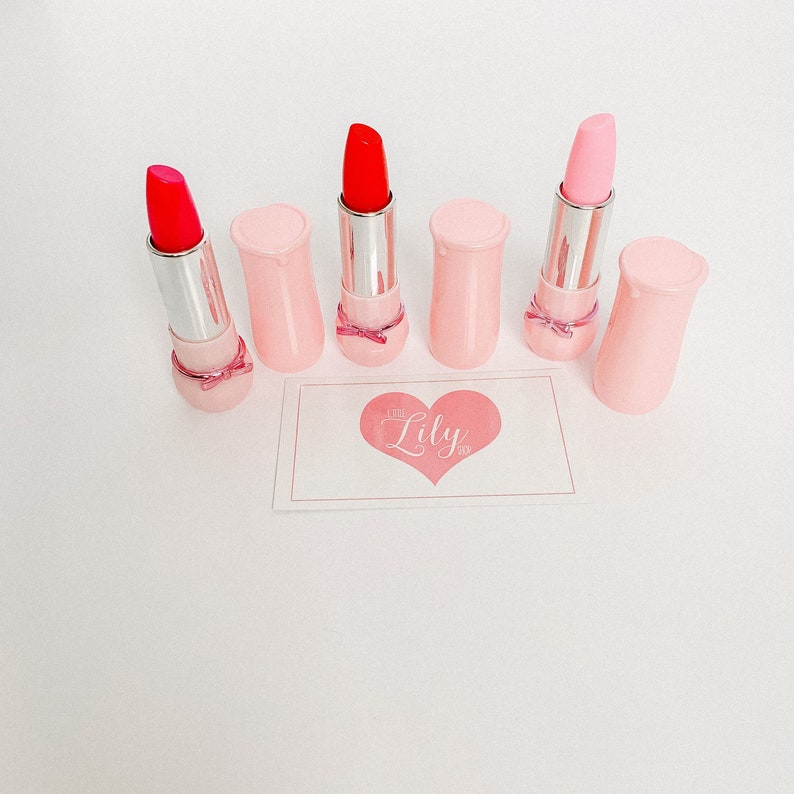 May include: Five pink lipstick tubes with a silver cap and a pink bow. The lipsticks are arranged in a row on a white surface. The text "You Lily" is printed on a pink heart-shaped card in the background.