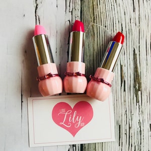 May include: Three pink lipstick tubes with silver caps and a pink bow detail. The lipsticks are in shades of pink, red, and light pink. The lipsticks are arranged on a white wooden surface with a white card that says "Little Lily Shop" in pink lettering.