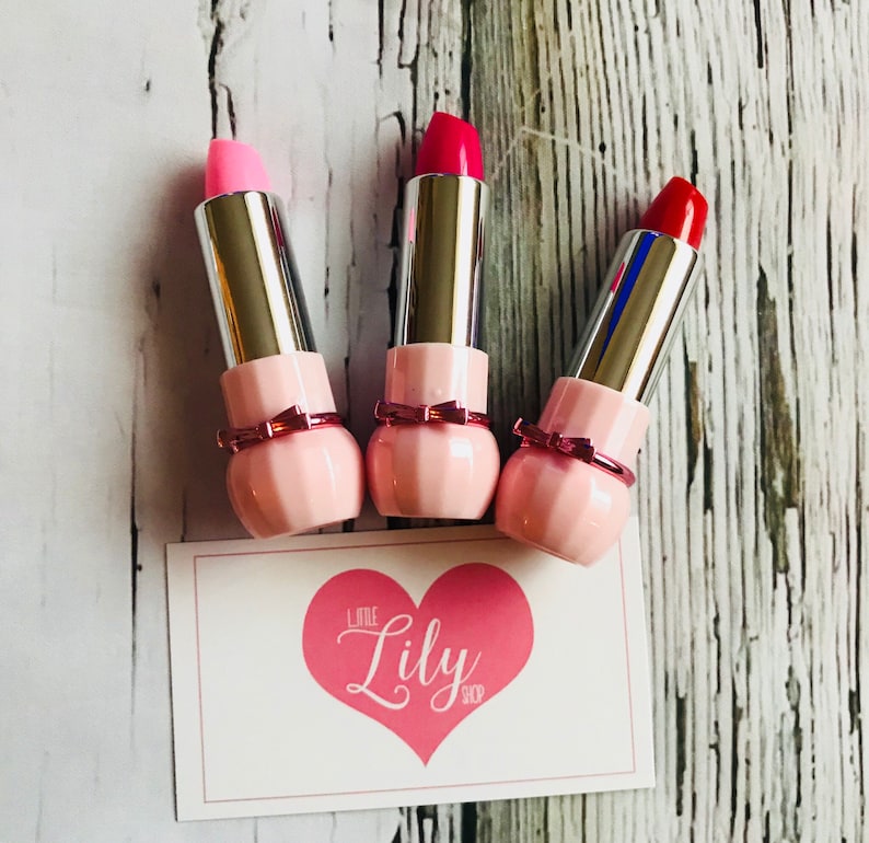 May include: Three pink lipstick tubes with silver caps and a pink bow detail. The lipsticks are in shades of pink, red, and light pink. The lipsticks are arranged on a white wooden surface with a white card that says "Little Lily Shop" in pink lettering.