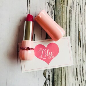 May include: A pink lipstick tube with a red lipstick bullet. The lipstick is in a pink tube with a bow detail. The tube is open and the lipstick is visible. The lipstick is on a white card with the text "Little Lily Shop" in pink.