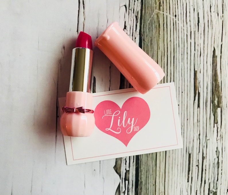 May include: A pink lipstick tube with a red lipstick bullet. The lipstick is in a pink tube with a bow detail. The tube is open and the lipstick is visible. The lipstick is on a white card with the text "Little Lily Shop" in pink.