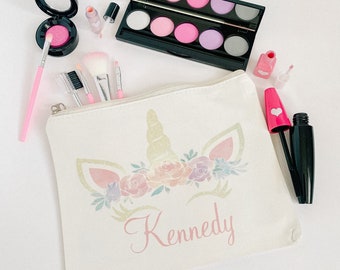 May include: A white canvas makeup bag with a unicorn design and the name "Kennedy" printed on it. The bag is filled with makeup items, including eyeshadow palettes, lip gloss, mascara, and brushes.