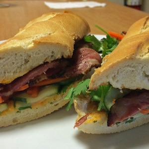 May include: Two halves of a Vietnamese sandwich, known as a banh mi, on a white plate. The sandwich is filled with meat, vegetables, and coriander. The bread is crusty and golden brown.