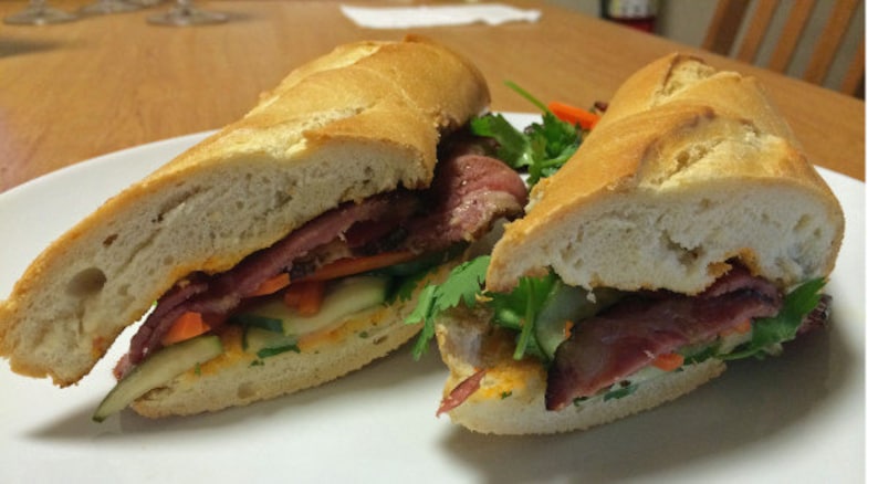 May include: Two halves of a Vietnamese sandwich, known as a banh mi, on a white plate. The sandwich is filled with meat, vegetables, and coriander. The bread is crusty and golden brown.