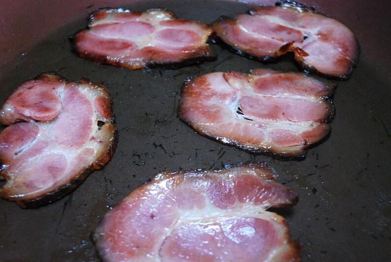 May include: Close-up of five rashers of cooked bacon in a frying pan. The bacon is crispy and has a light brown colour.