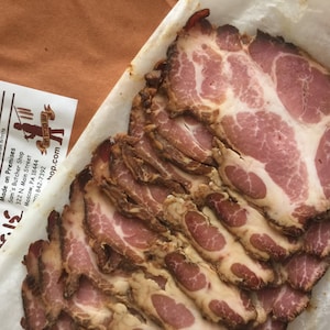 May include: Close-up of thinly sliced, smoked buckboard bacon. The bacon is arranged in a stack on white parchment paper. The label reads "Buckboard Bacon" and lists the ingredients as pork, salt, spices, and sodium nitrite. It also states that the bacon is "Made on Premises" at Sam's Butcher Shop in Moscow, PA.