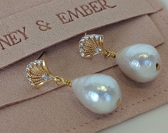 May include: A pair of gold-tone drop earrings featuring a fan-shaped top with small clear stones and a large white teardrop-shaped pearl. The earrings are displayed on a light pink suede pouch with the text HONEY & EMBER embossed on the front flap.