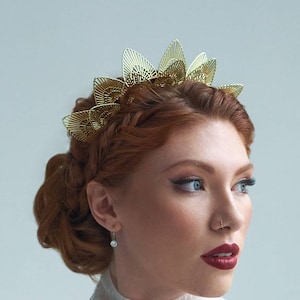 May include: A woman with red hair wearing a gold crown headband and a white lace top. She is looking to the right of the frame.