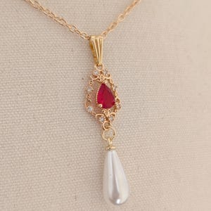 Puede incluir: Collar dorado con un colgante en forma de lágrima con una piedra preciosa roja y pequeñas piedras transparentes. Una perla blanca en forma de pera cuelga debajo. La cadena es delicada y el colgante tiene un diseño ornamentado.