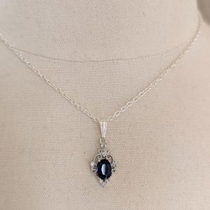 Può includere: Una collana d'argento con un pendente con una grande gemma ovale blu scuro. La gemma è circondata da pietre più piccole e trasparenti in un'incastonatura decorativa. La collana è esposta su uno sfondo di tessuto neutro.