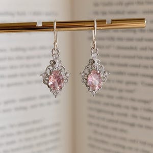 Puede incluir: Un par de pendientes colgantes plateados. Cada pendiente presenta una piedra preciosa rosa de talla ovalada engastada en un marco decorativo calado. El marco está adornado con piedras preciosas más pequeñas y transparentes, y los pendientes cuelgan de un gancho simple.
