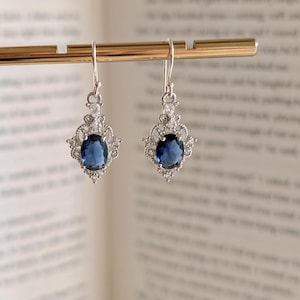 Puede incluir: Un par de pendientes colgantes plateados con una piedra preciosa ovalada azul oscuro. Los pendientes tienen un diseño de filigrana decorativo alrededor de la piedra preciosa.