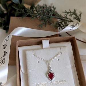 May include: A silver necklace with a red gemstone pendant, displayed in a gift box. The pendant is surrounded by small, clear stones. The box is brown with a white interior, and a white ribbon with the words "Honey & Ember" is wrapped around it.