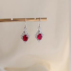 May include: A pair of silver drop earrings, each with a central, oval, deep red gemstone. The red stone is encircled by smaller, clear gemstones. The earrings hang from a simple hook.