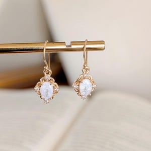 Puede incluir: Un par de pendientes de aro dorados con una piedra ovalada blanca engastada en un marco decorativo. Los pendientes tienen un diseño delicado de inspiración vintage.