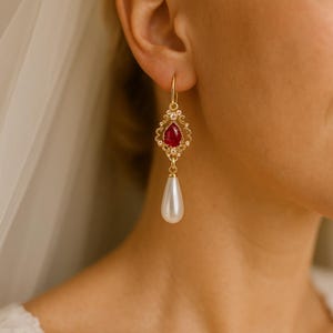 Puede incluir: Pendientes colgantes dorados con una gema roja y una perla blanca en forma de lágrima. Los pendientes tienen un diseño ornamental de filigrana con pequeñas piedras transparentes. Los pendientes se muestran en una oreja.