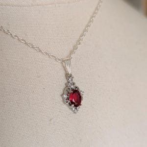 May include: Silver necklace with a pendant featuring a large, oval, deep red gemstone. The pendant is surrounded by smaller, clear gemstones. The necklace is displayed on a neutral background.