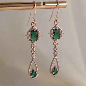 May include: A pair of gold-coloured drop earrings. Each earring features a rectangular emerald-green gemstone at the top, surrounded by small clear stones. A teardrop-shaped emerald-green stone dangles below. The earrings are suspended from a gold-coloured hook.