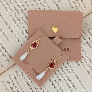 May include: Gold earrings featuring a red, teardrop-shaped gemstone and a white pearl drop. The earrings are presented on a pink card with the text "HONEY & EMBER". A small pink pouch with a gold button is also visible.