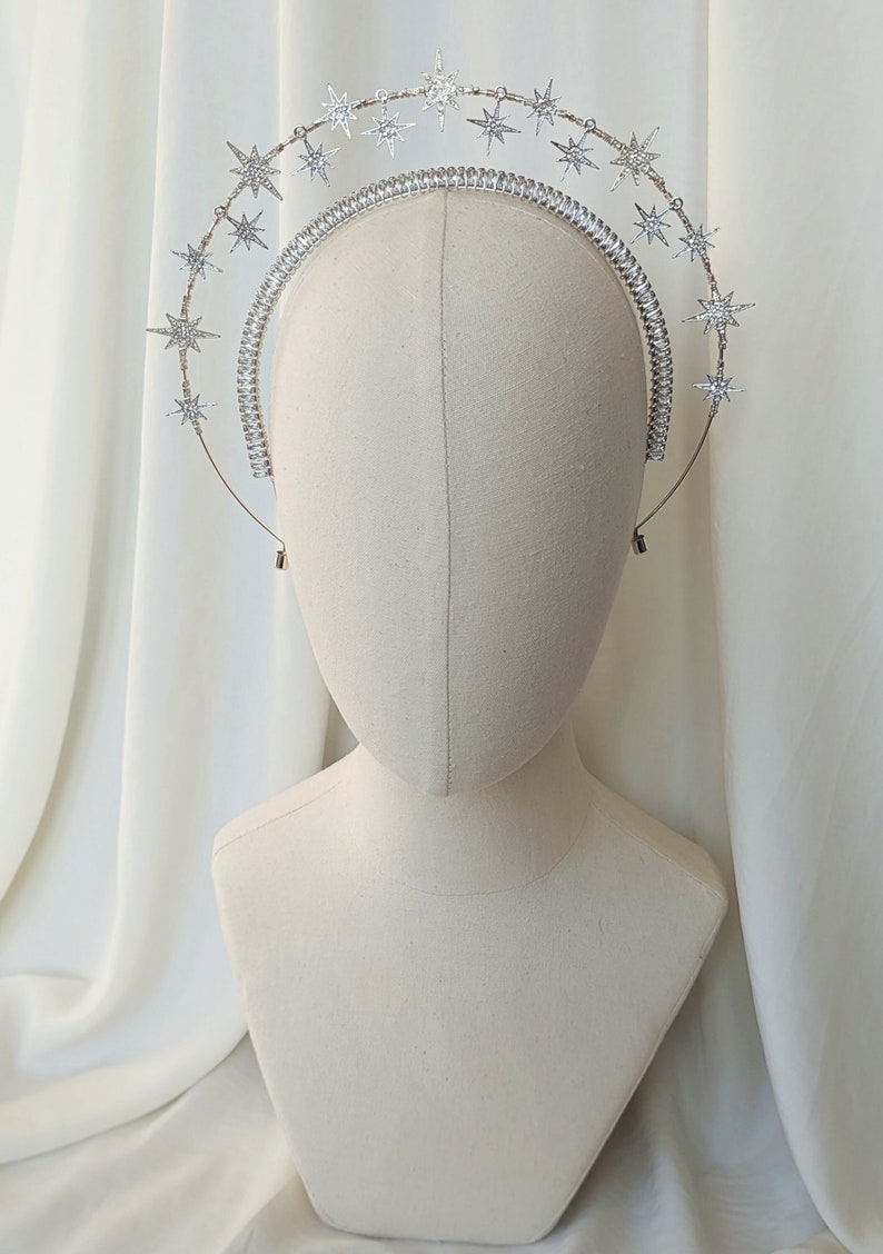 Puede incluir: Diadema plateada con una doble fila de piedras de imitaci&oacute;n en forma de estrella. La diadema est&aacute; en una cabeza de maniqu&iacute; blanca.