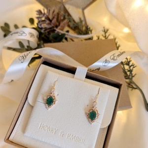 May include: Gold earrings featuring emerald-green oval gemstones and small clear stones. The earrings are presented in a white box with the text "HONEY & EMBER". The box is inside a brown gift box, tied with a white ribbon, ideal for gifting.