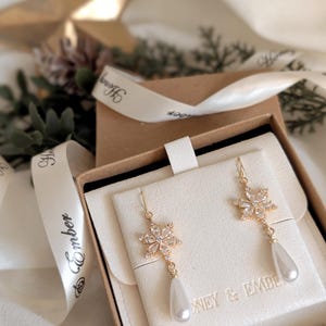 Pode incluir: Brincos dourados com design floral e pingentes de pérolas em forma de gota, apresentados numa caixa de presente. Os brincos apresentam cristais transparentes e são exibidos em um fundo creme. A caixa é amarrada com uma fita que diz "Honey & Ember."