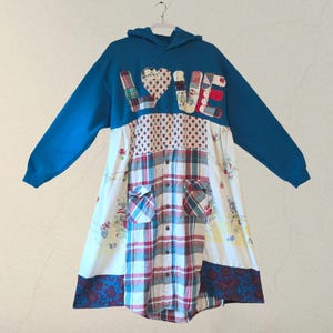Puede incluir: Un vestido azul con capucha y mangas largas. La palabra "LOVE" está aplicada en el pecho con tela de patchwork. El vestido presenta una mezcla de telas florales, a cuadros y estampadas.