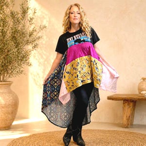 May include: A woman wearing a black t-shirt with text and a patchwork tunic. The tunic features panels of pink, yellow floral, and patterned fabric, with a high-low hemline. She is also wearing black leggings and black boots.