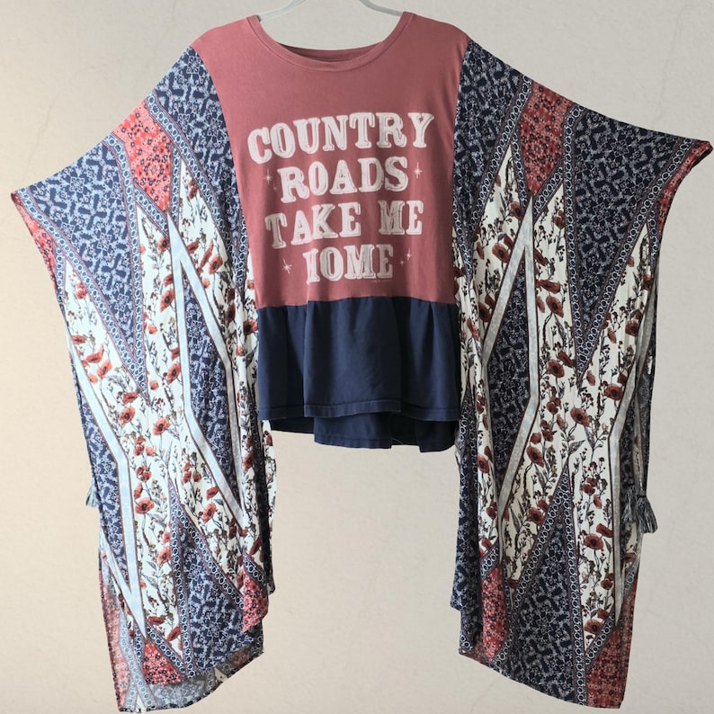 Pu&ograve; includere: Top rosso e blu navy con la scritta bianca "COUNTRY ROADS TAKE ME HOME". Il top ha maniche fantasia con disegni floreali e geometrici rossi, bianchi e blu. Il top &egrave; appeso a una gruccia bianca.