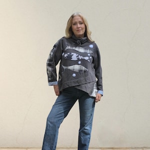 Peut inclure: Un sweat-shirt gris foncé avec trois applications de poissons en motif écossais. Le sweat-shirt a un design asymétrique unique avec des accents écossais sur les manches et un col montant. Le modèle porte un jean bleu.