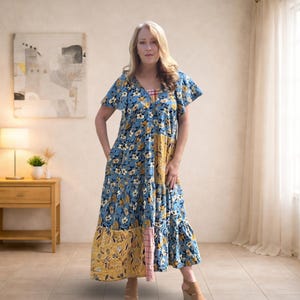 May include: A blue floral print maxi dress with short sleeves and a V-neck. The dress features a patchwork design with yellow and pink accents. The model is wearing the dress in a well-lit room with a neutral background.