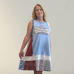 May include: A sleeveless dress made from repurposed t-shirts. The top is light blue with a "Bubba Gump Shrimp Co." logo. The skirt features patchwork of red, white, floral, and gray fabrics. The dress is a unique, casual style.