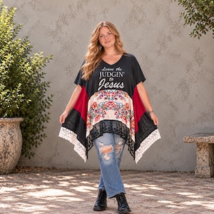 May include: A black short-sleeved poncho-style top featuring the text "Leave the JUDGIN' to Jesus" in white. The garment includes a central cream-coloured panel with a colourful floral print, red side panels, and a black lace hem with white trim.