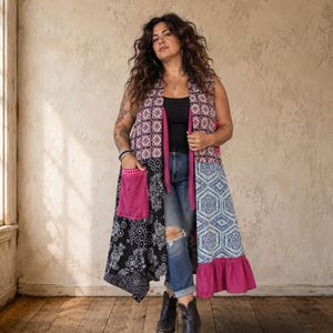 May include: A sleeveless long vest featuring a patchwork design with various patterns including black and white paisley, geometric blue and white prints, and pink accents. The garment includes a solid pink pocket and a ruffled pink hemline.