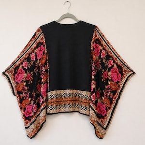 May include: A black top with wide, flowing sleeves featuring a vibrant floral pattern in shades of pink, orange, and cream. The garment includes a decorative geometric border along the hem and sleeve edges, set against a solid black center panel.