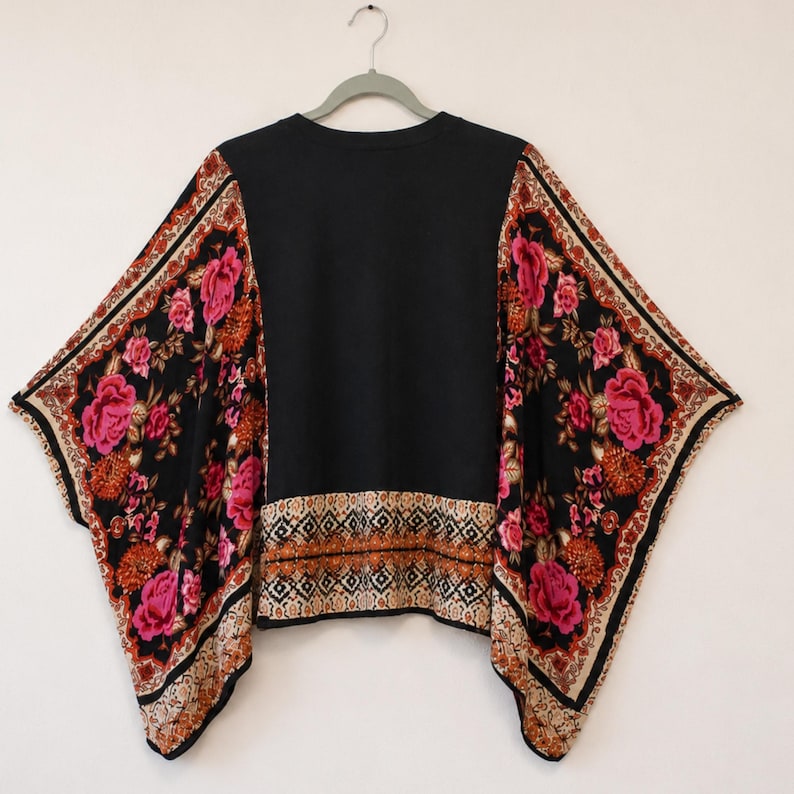 May include: A black top with wide, flowing sleeves featuring a vibrant floral pattern in shades of pink, orange, and cream. The garment includes a decorative geometric border along the hem and sleeve edges, set against a solid black center panel.