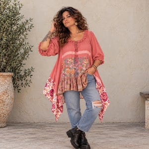 Könnte beinhalten: Eine altrosa Tunika mit gerüschtem Ausschnitt, Dreiviertelärmeln und einem floral gemusterten Mittelteil. Der Saum ist asymmetrisch und mit einer dekorativen Blumenborte versehen. Die Tunika wird mit einer hellblauen, zerrissenen Jeans kombiniert.