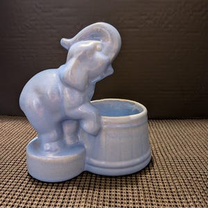 May include: A light blue ceramic elephant planter. The elephant is standing next to a round planter. The elephant has its trunk raised. The planter is a light blue color.