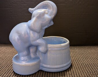Puede incluir: Una maceta de elefante de cerámica azul claro. El elefante está de pie junto a una maceta redonda. El elefante tiene la trompa levantada. La maceta es de color azul claro.