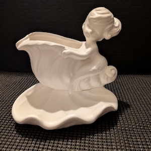 Puede incluir: Una maceta de cerámica blanca con forma de sirena. La figura de la sirena está esculpida con cabello suelto y un vestido delicado, sosteniendo una gran concha abierta. La concha sirve como maceta, y la base tiene forma de concha a juego.