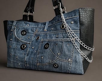 May include: A tote bag constructed from blue denim fabric featuring black leather-like side panels with a crocodile texture. The denim section is embellished with silver-tone metal grommets, studs, and a silver chain strap. It has black handles.